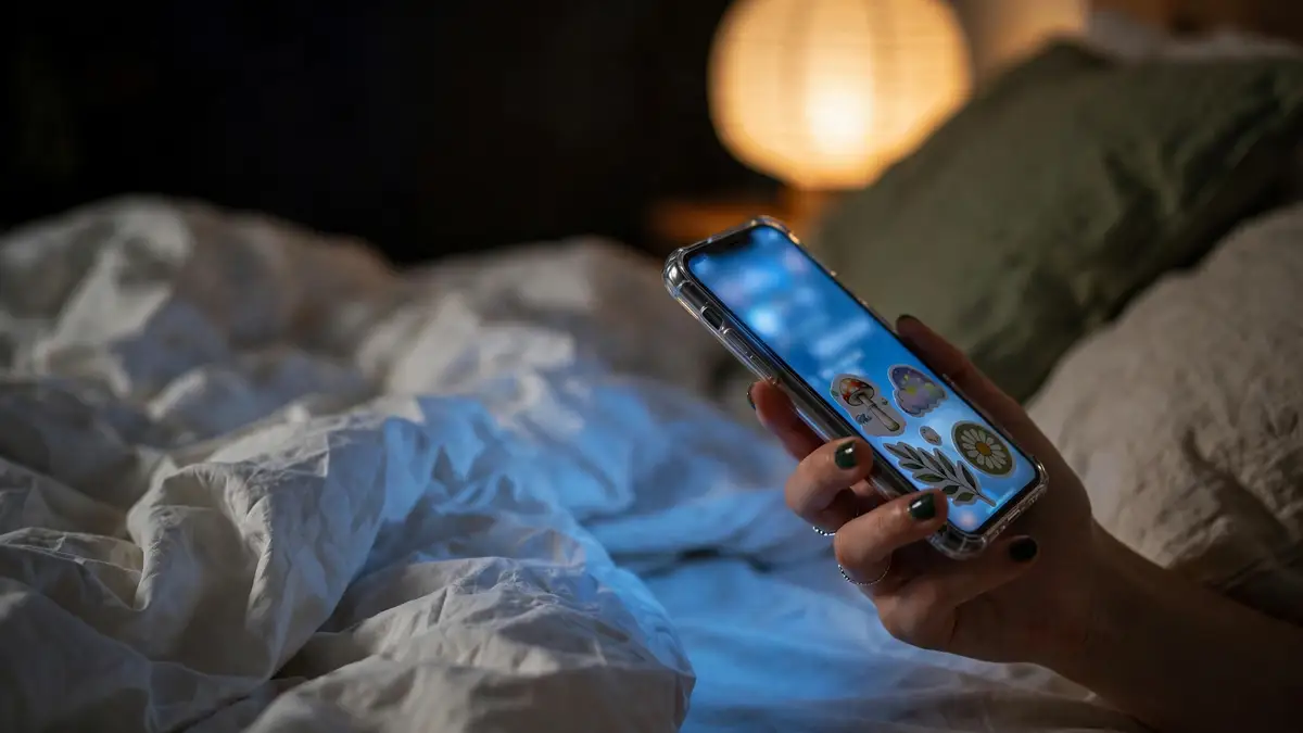Una mano con las uñas pintadas de verde sostiene de noche un móvil con la pantalla encendida, sobre una cama deshecha